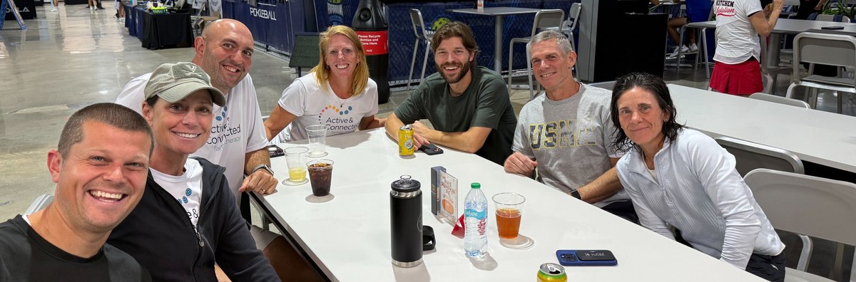 Seven pickleball friends and partners gathered around a table at a tournament venue, smiling together between matches