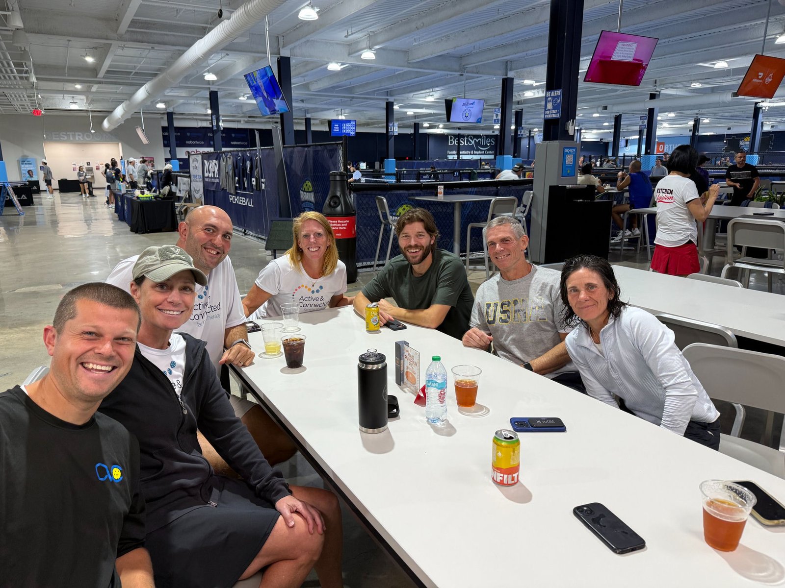 Seven pickleball friends and partners gathered around a table at a tournament venue, smiling together between matches
