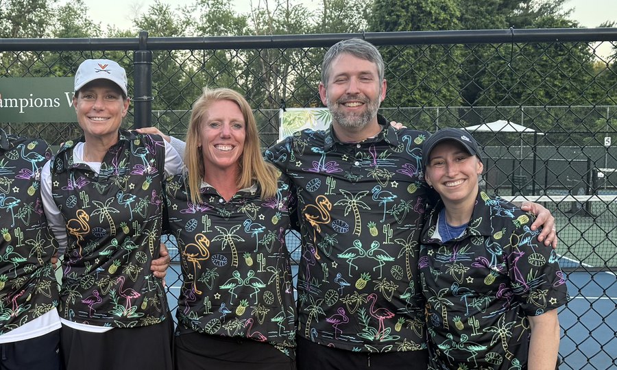 Four pickleball teammates in matching flamingo-print jerseys, arms around each other by the court fence at dusk