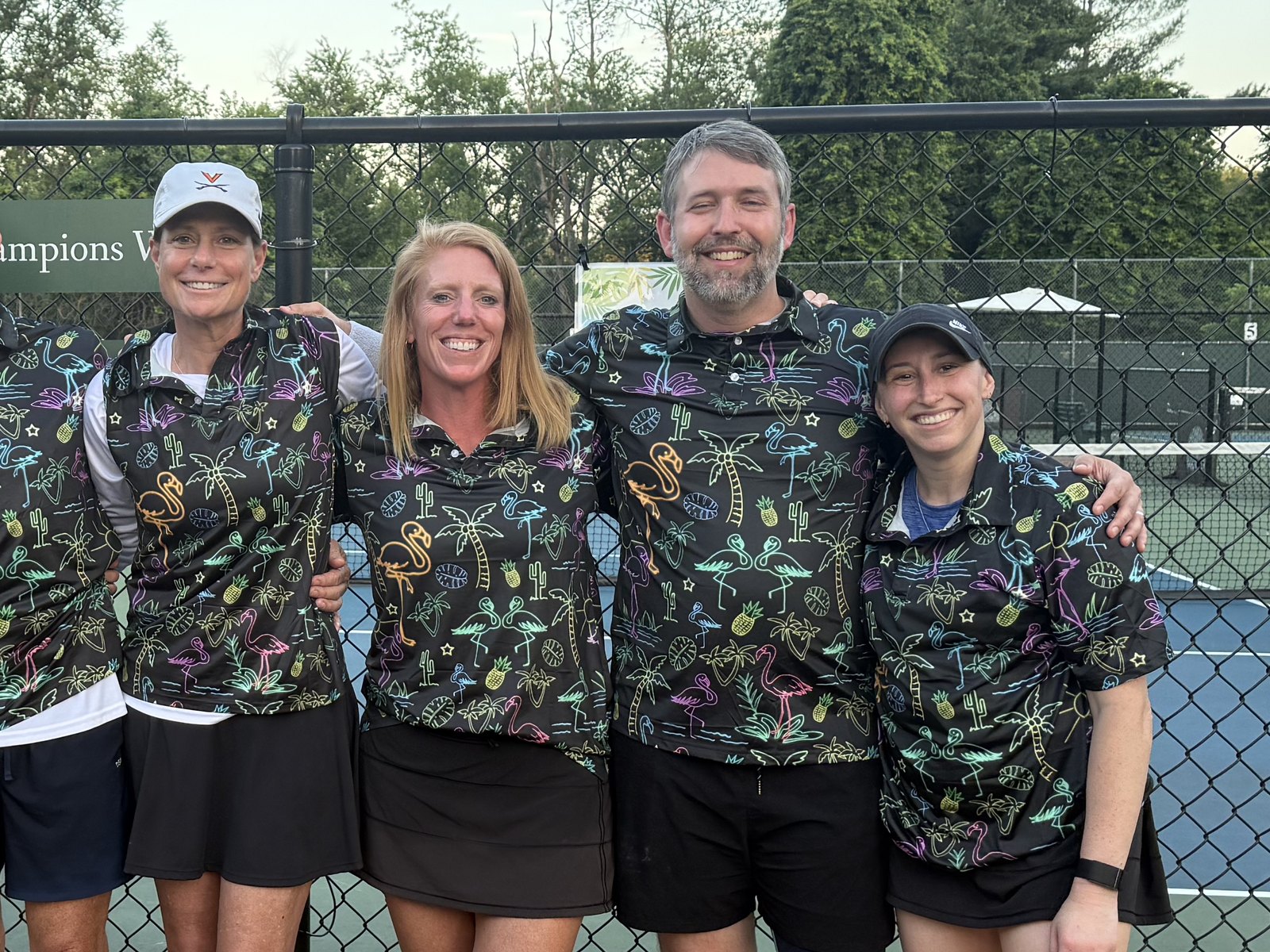 Four pickleball teammates in matching flamingo-print jerseys, arms around each other by the court fence at dusk