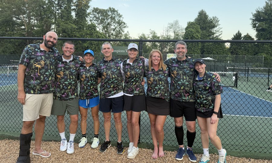 A pickleball team in matching flamingo-print jerseys gathered together by the court fence after a match