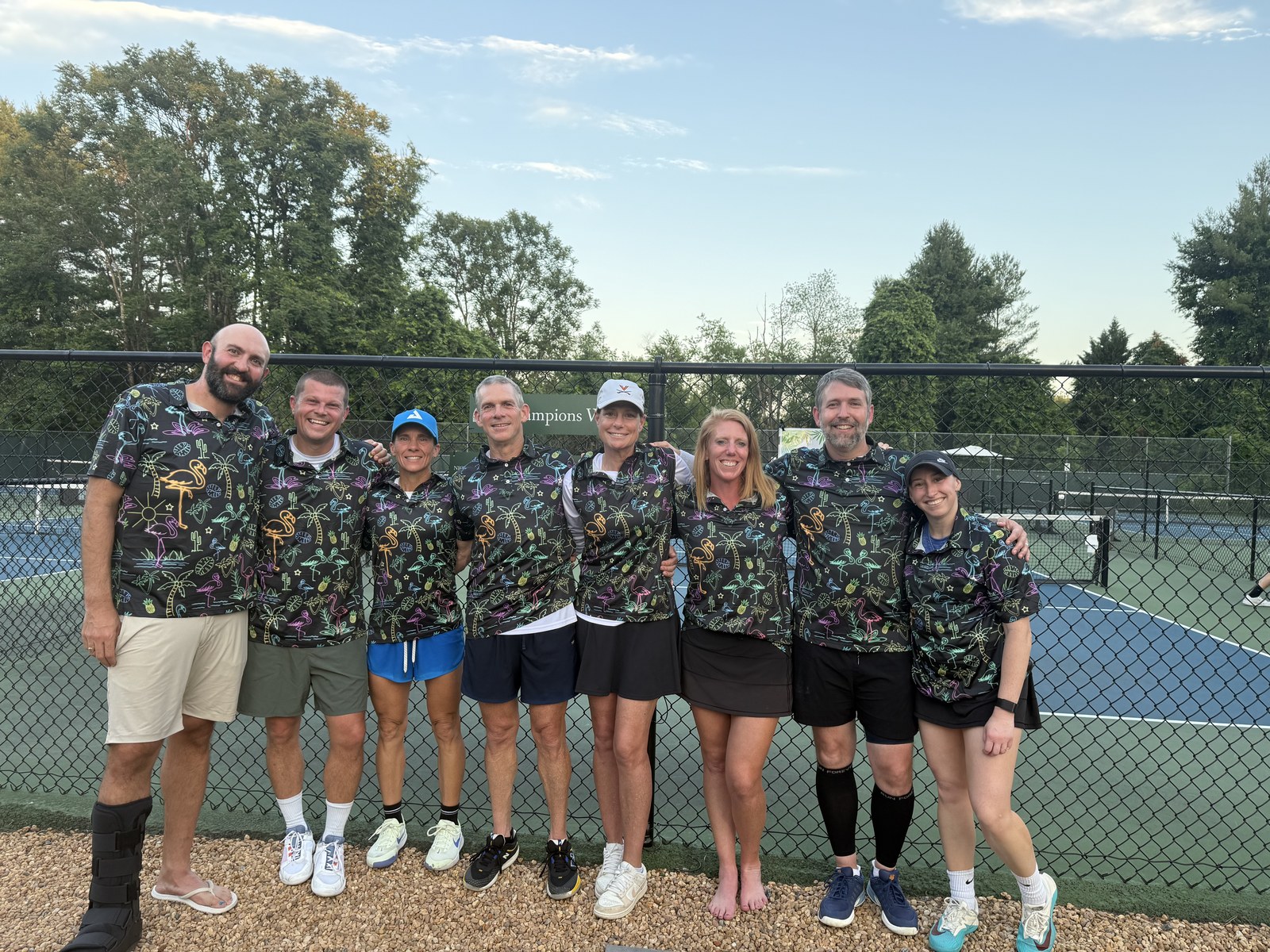 A pickleball team in matching flamingo-print jerseys gathered together by the court fence after a match